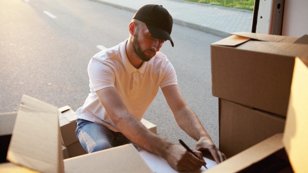 A delivery man in a black cap writing something on a piece of paper at the back of his delivery van